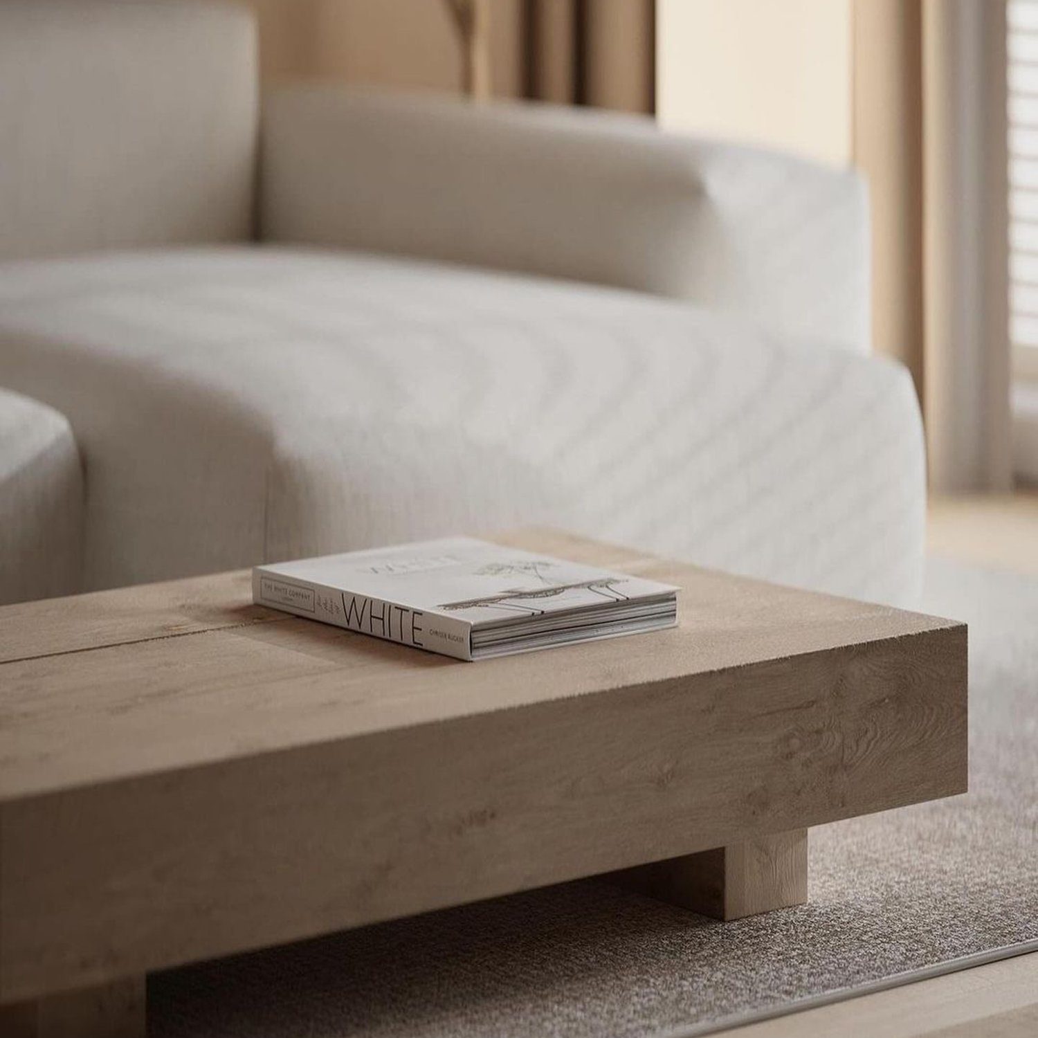 A minimalist living room featuring a low-profile wooden coffee table with a book titled "WHITE" on top, in front of a softly textured off-white sofa