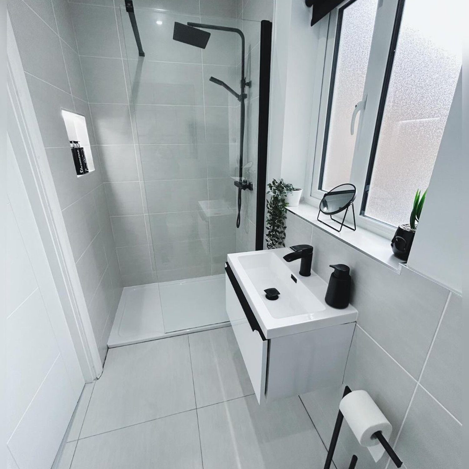 A sleek and modern designed monochromatic bathroom featuring grey tiles, stylish matte black fixtures, a wall-mounted sink with undershelf, and a clear glass shower partition.