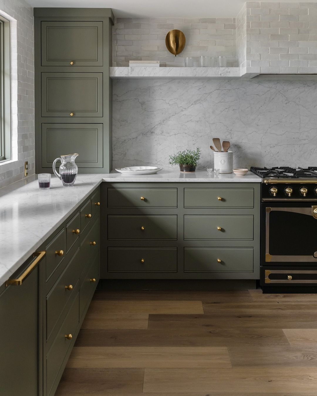 A sophisticated modern kitchen featuring olive green cabinetry, brass hardware, marble countertops, and a classic black range cooker.