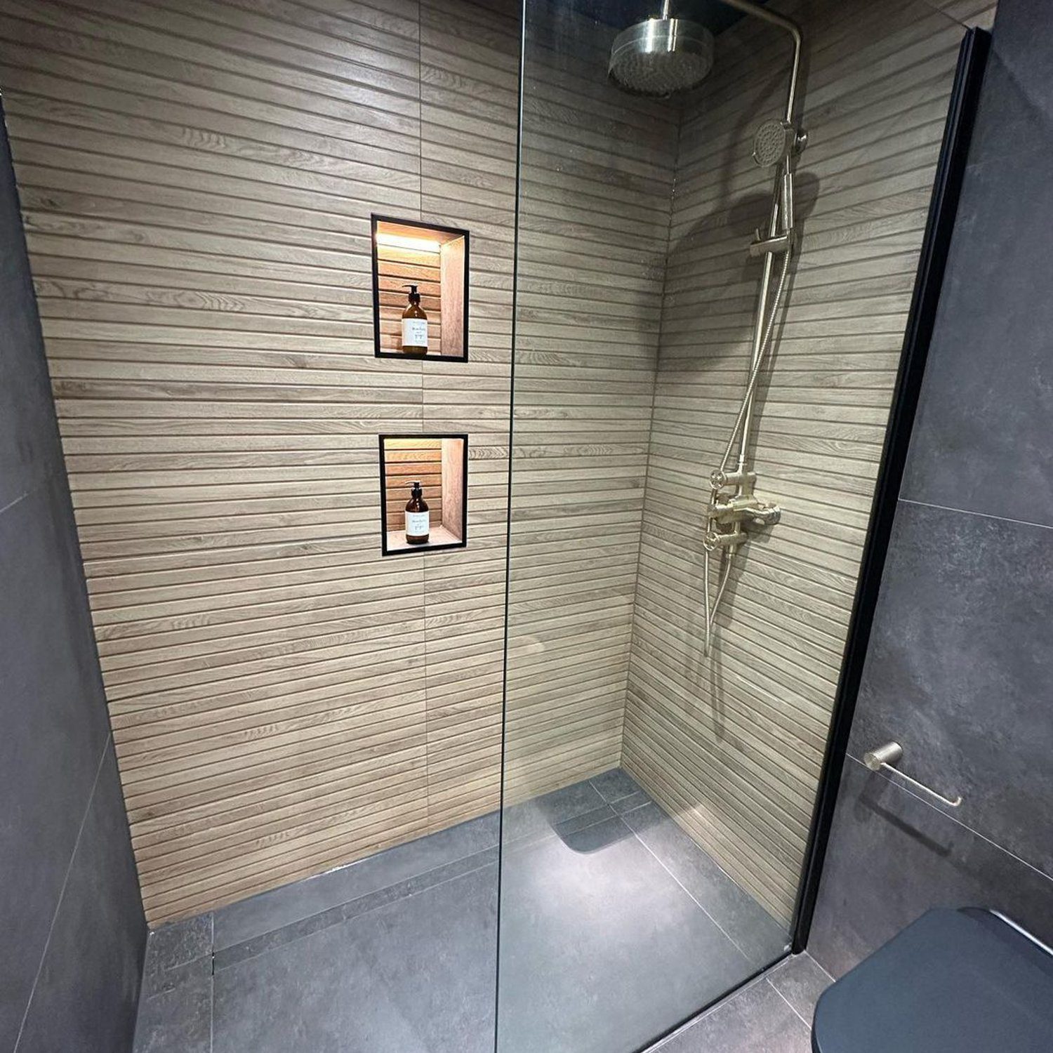 A modern shower space with beige textured wall tiles, glass partition, and stainless steel fixtures