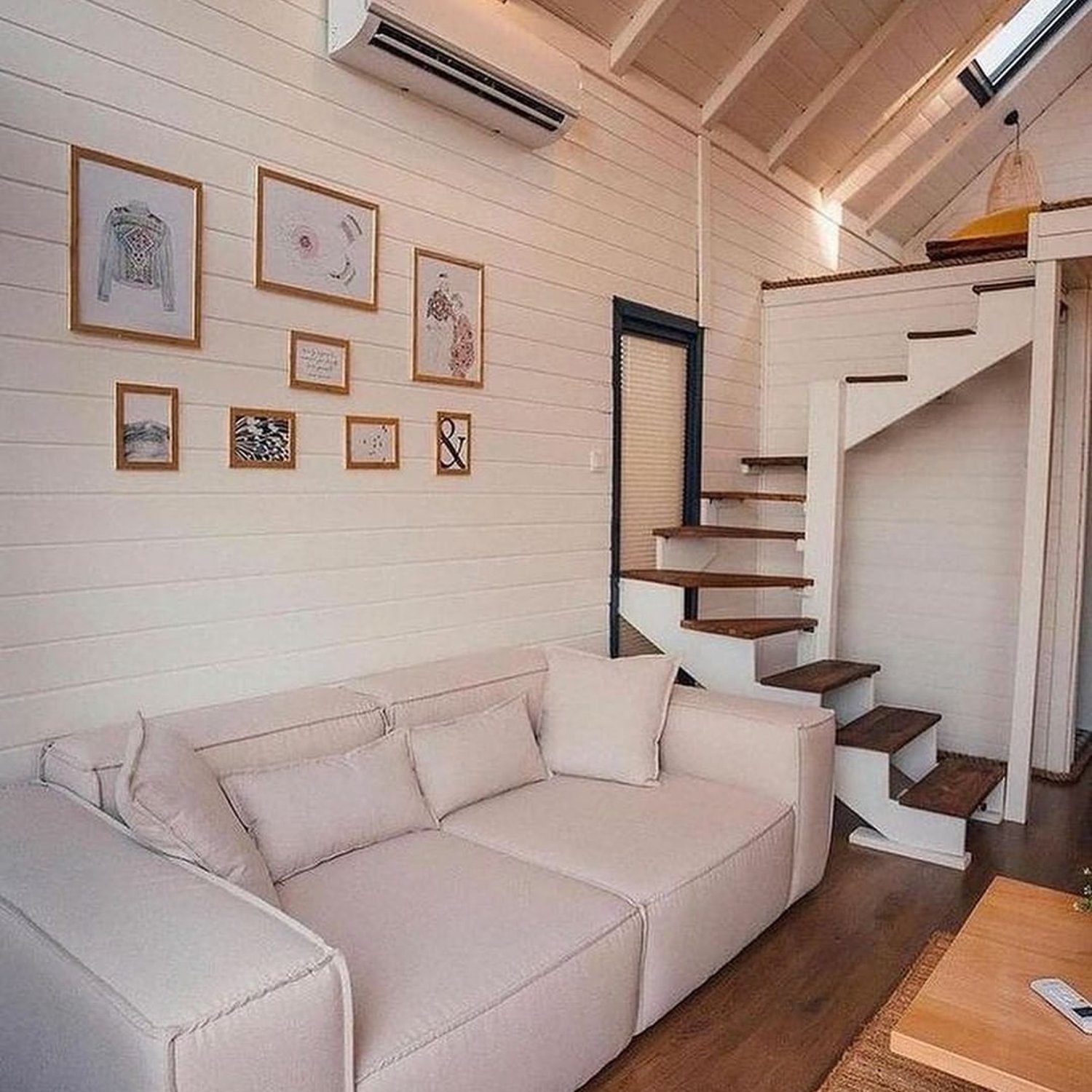 A cozy and modern tiny house interior featuring a minimalist aesthetic with a light beige sectional sofa and floating wooden stairs leading to a loft space.