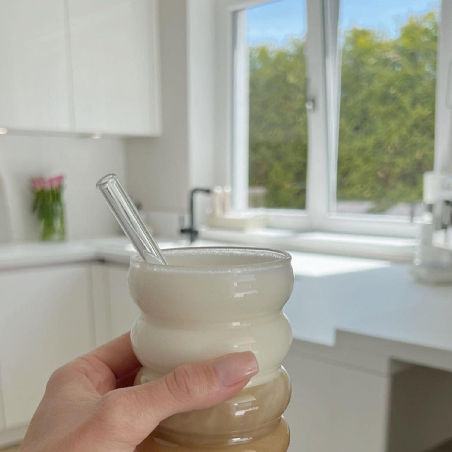 A person holding a layered coffee drink in a glass with a glass straw, with a bright kitchen and window view in the background