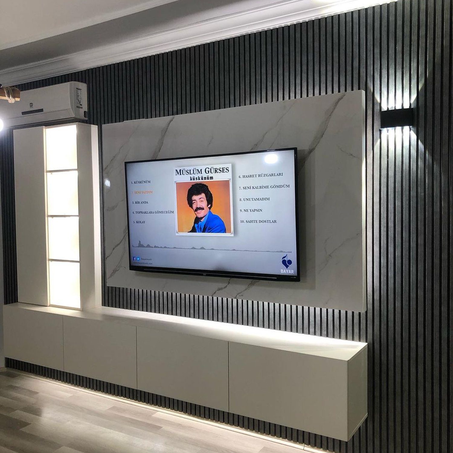 A modern interior display featuring a large television screen with an image of Müslüm Gürses set into a marble wall, complemented by vertical black stripes and a white cabinet below