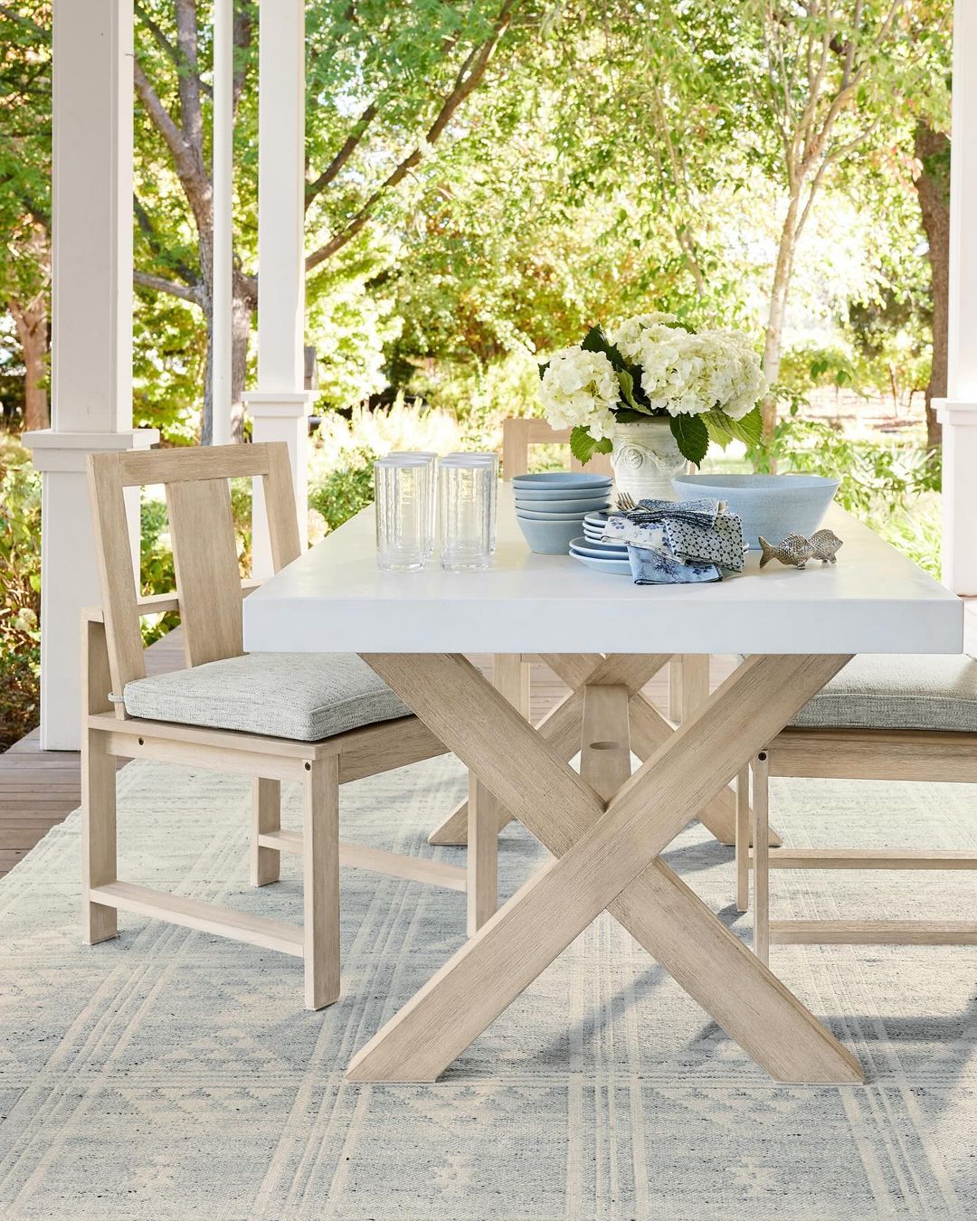 An outdoor dining area with a white tabletop and a crossed wooden base surrounded by matching chairs on a soft blue patterned rug, set against a backdrop of greenery.