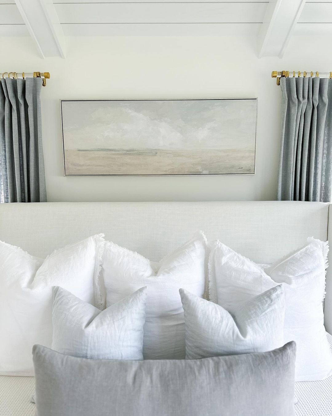 A serene bedroom setting focusing on a plush, white sofa adorned with soft pillows against a backdrop of a landscape painting and stylish gray curtains