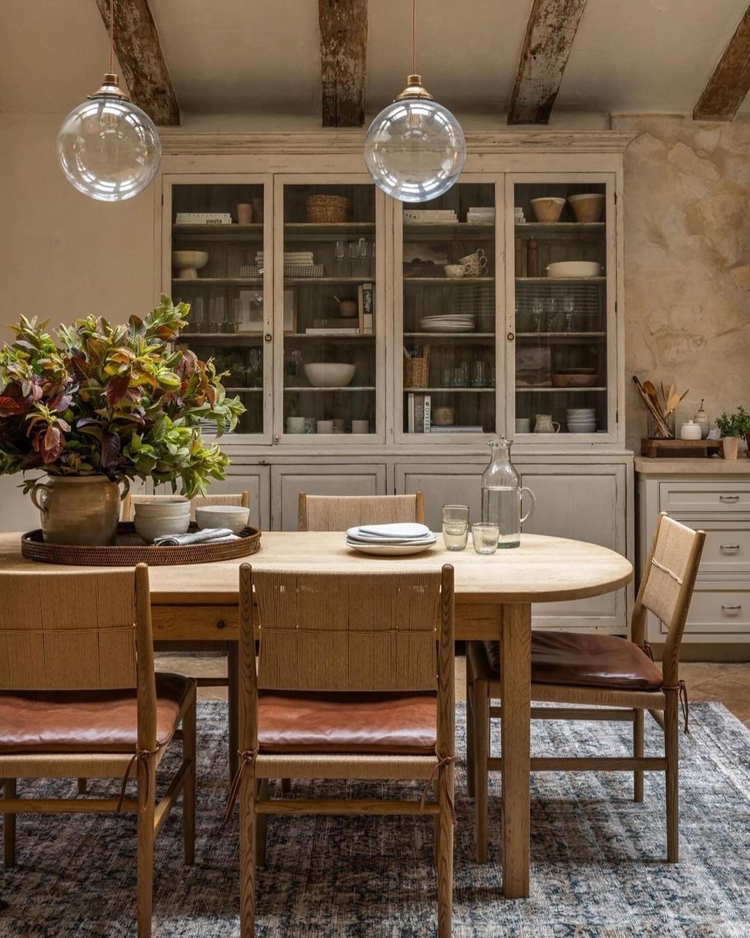 A rustic-style kitchen featuring a wooden table with terracotta-colored leather chairs, exposed wooden beams above, and a vintage glass-fronted cabinet filled with crockery.