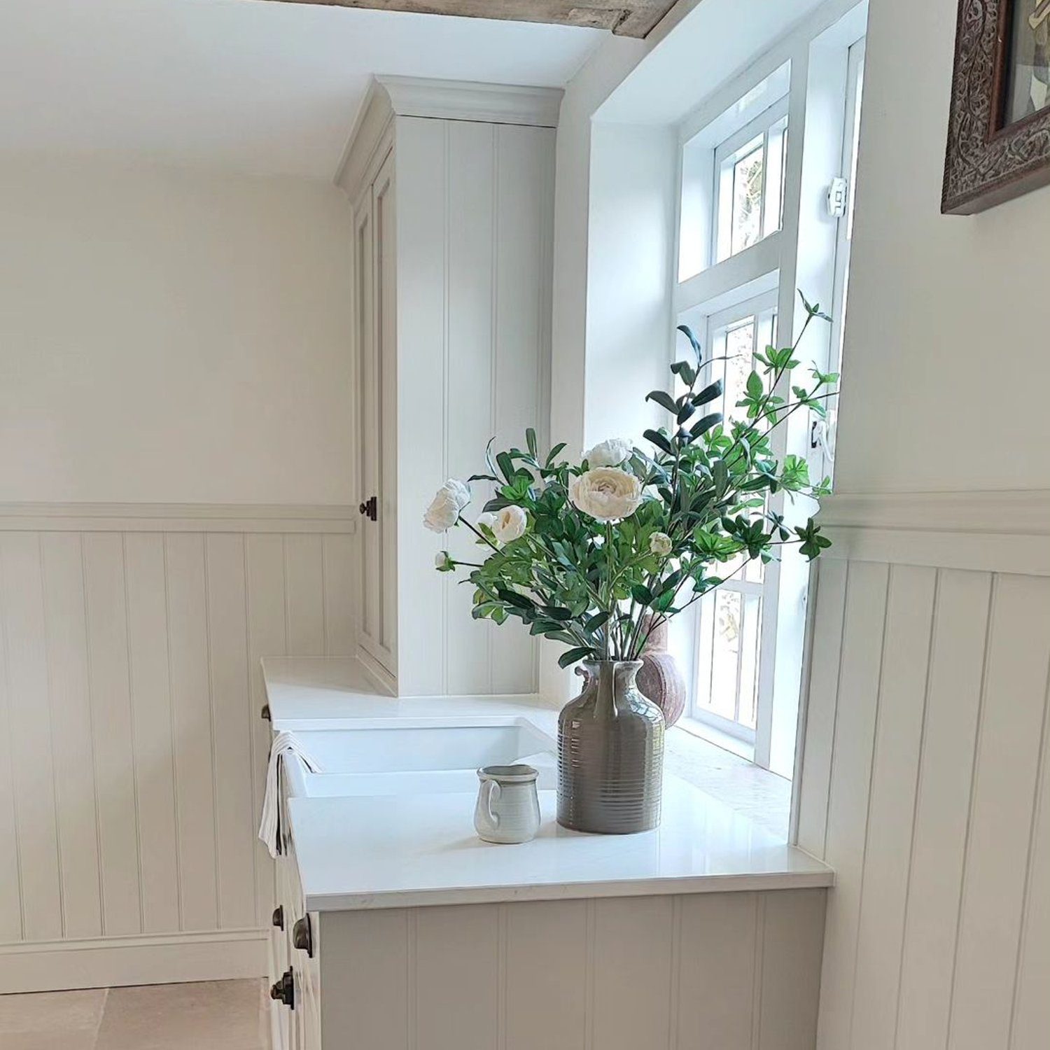 A quaint and charming corner featuring a farmhouse-style sink with beadboard paneling, set against a rustic wooden beam and bright window with a potted vase of fresh white roses placed on the countertop.