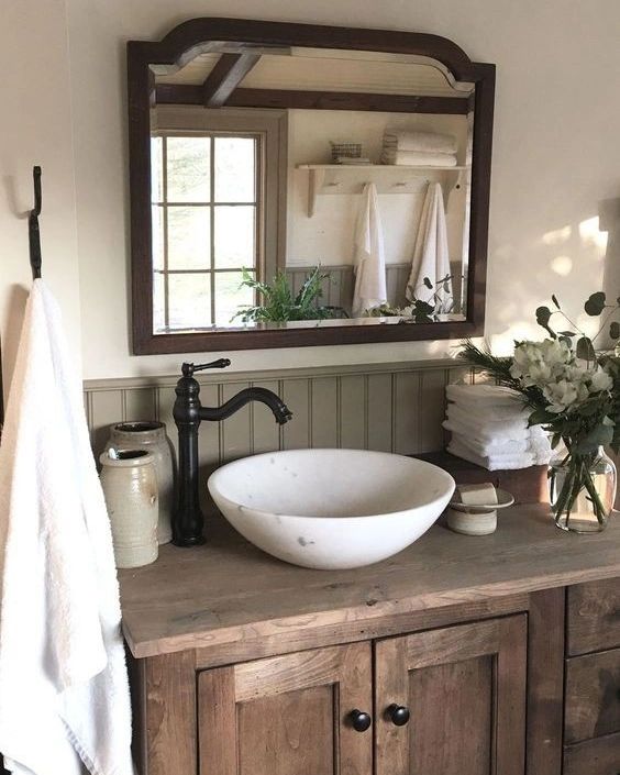 A rustic farmhouse-style bathroom featuring a wooden vanity with paneled drawers and black hardware, topped by a white oval vessel sink beneath a large wood-framed mirror.