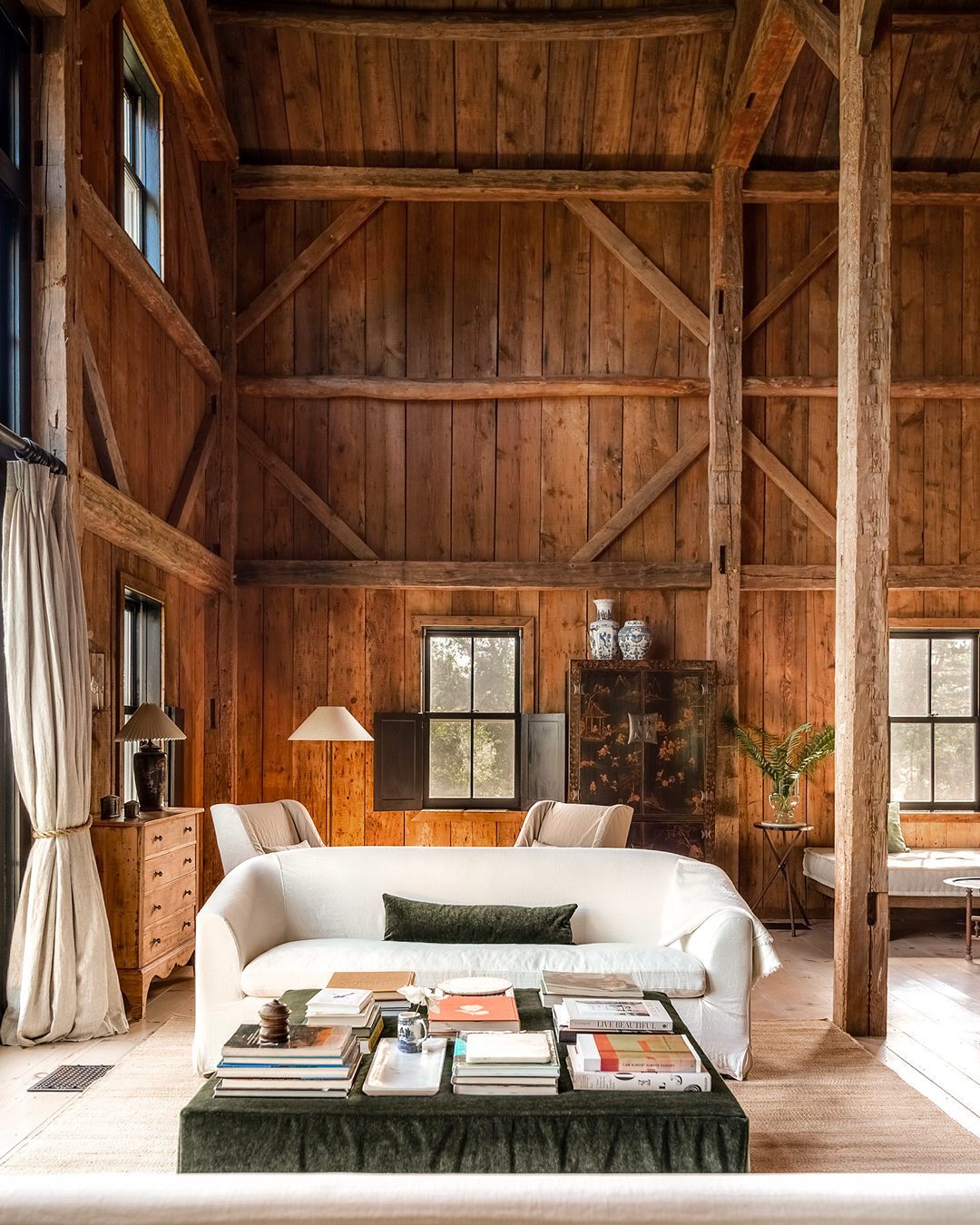 A cozy and rustic living room area with wooden walls and beams, large windows and a plush green ottoman