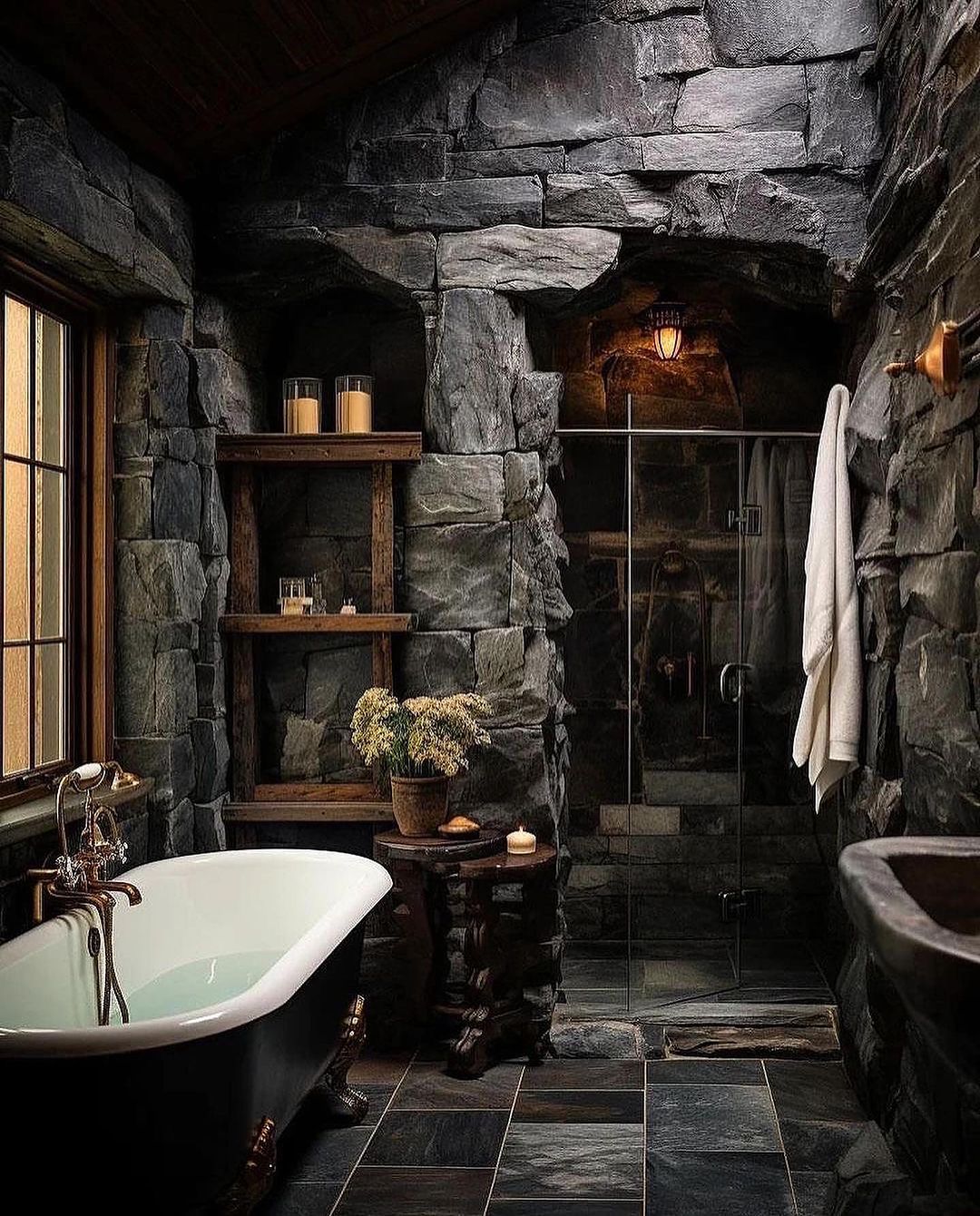 A rustic stone-clad bathroom with a claw-foot tub, glass-walled shower, and wooden accents.