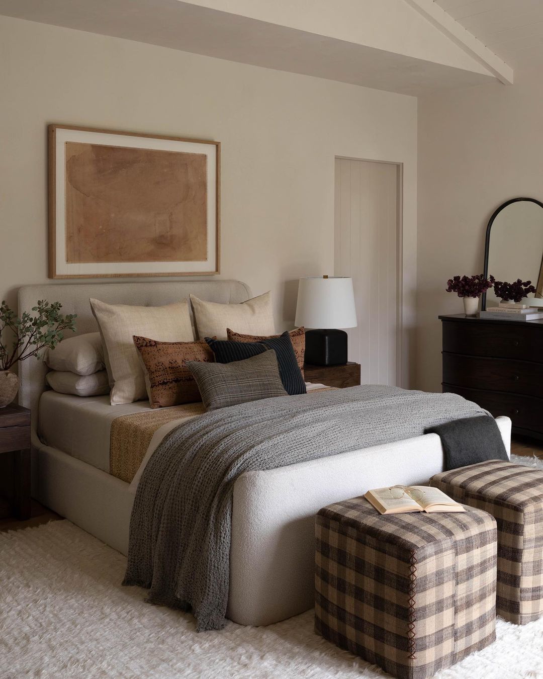 A tastefully appointed bedroom featuring a plush beige bed with a mix of textured pillows, a cozy gray knit throw blanket, and checkered ottomans at the foot