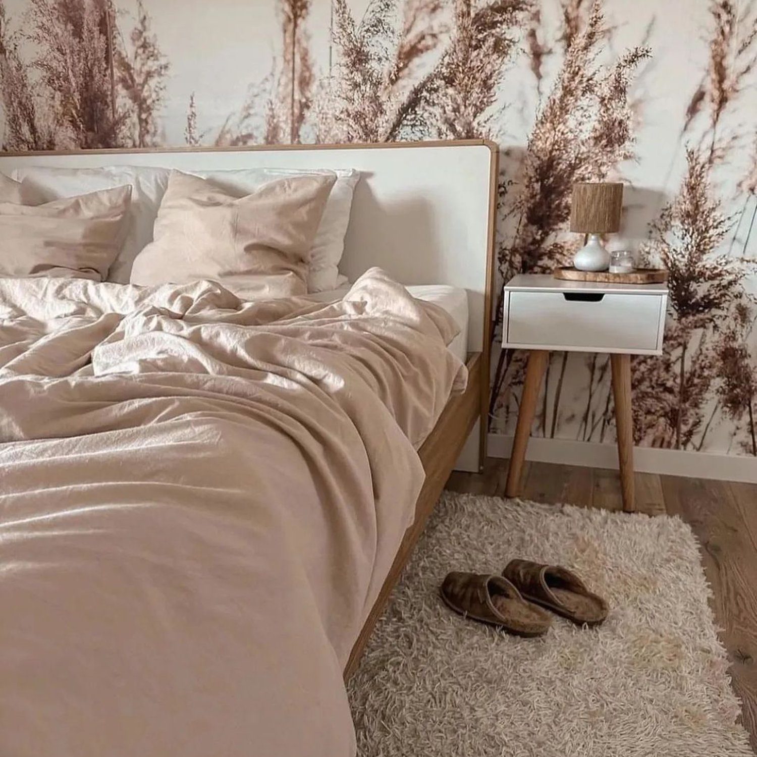 A serene bedroom featuring a blush satin bedding set, against a backdrop of tall pampas grass wallpaper, accompanied by a modern nightstand with a wood-and-ceramic lamp, positioned above a plush cream rug with a pair of cozy slippers nestled on it.