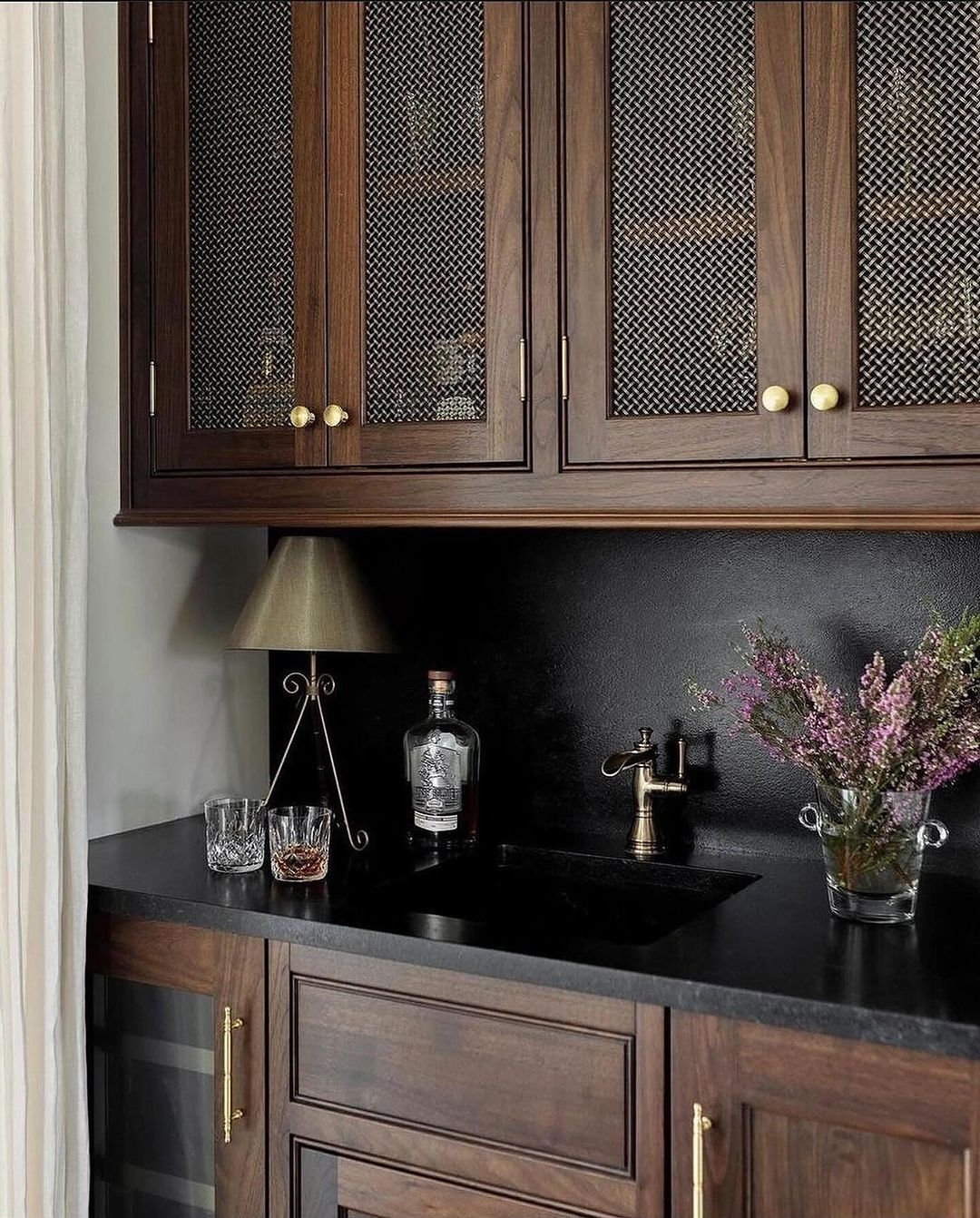 A sophisticated home bar area featuring dark wood cabinetry with mesh inserts, brass hardware, black countertop, and accent lighting