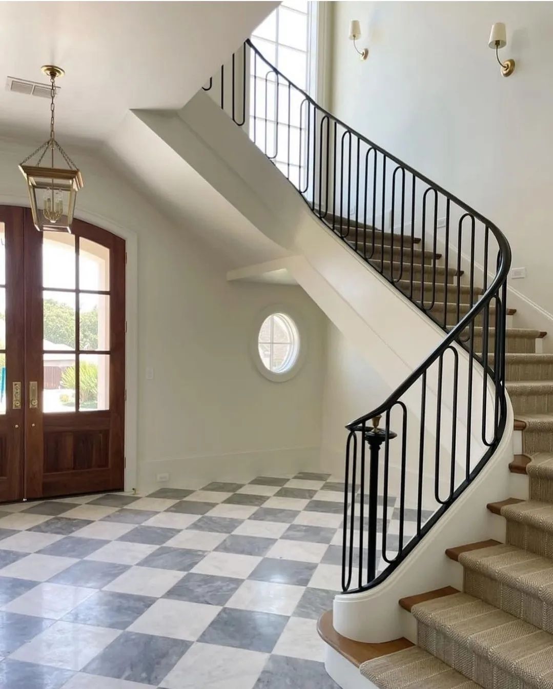 A spacious foyer featuring a winding staircase with a black wrought-iron railing, checkered floor tiles, and a round window