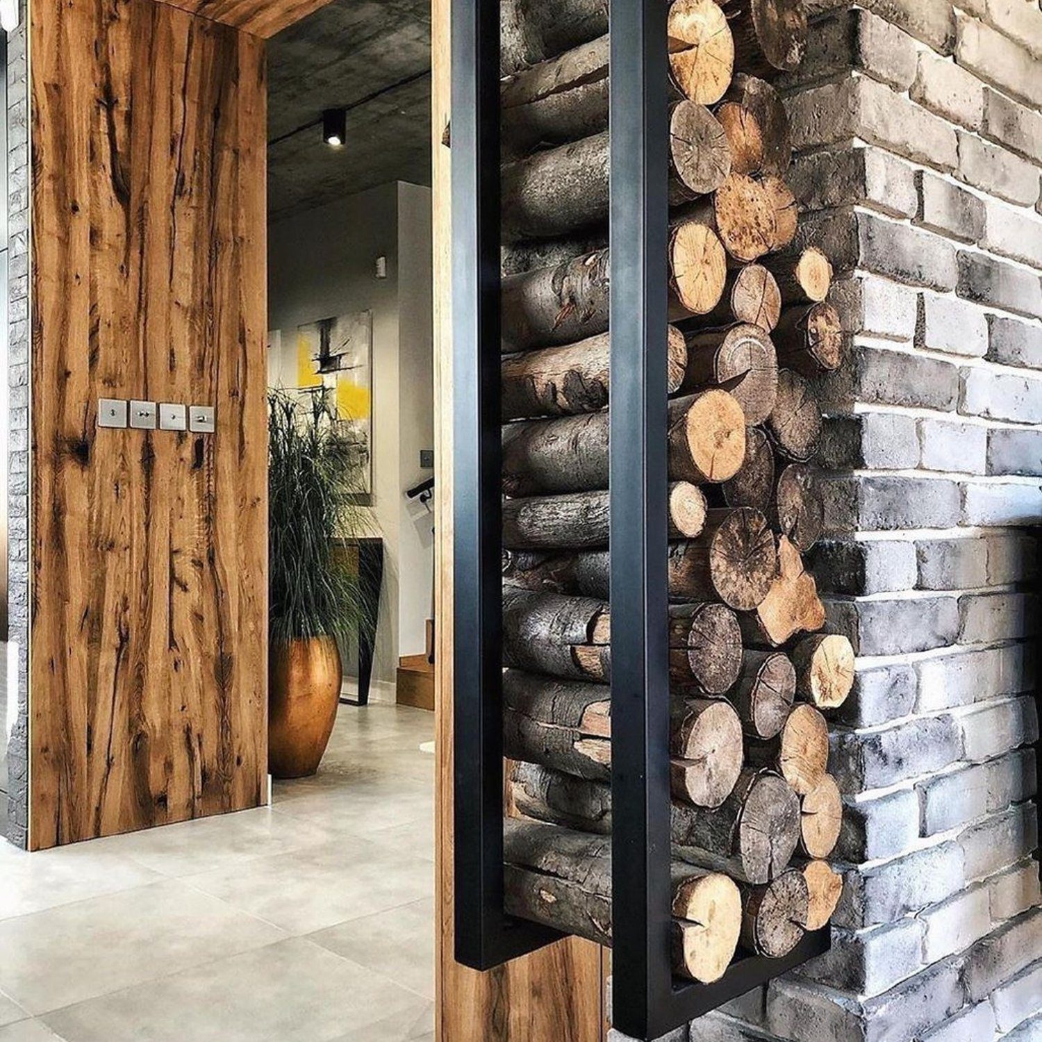 A harmoniously designed interior space featuring a rustic wood door frame, a brick column with neatly stacked firewood, and a peek into a modern room with chic decor.