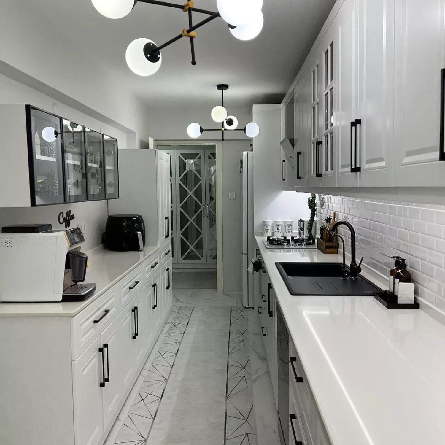 A modern, narrow kitchen with white cabinetry and subway tile backsplash, featuring geometric patterned floor tiles and stylish spherical light fixtures