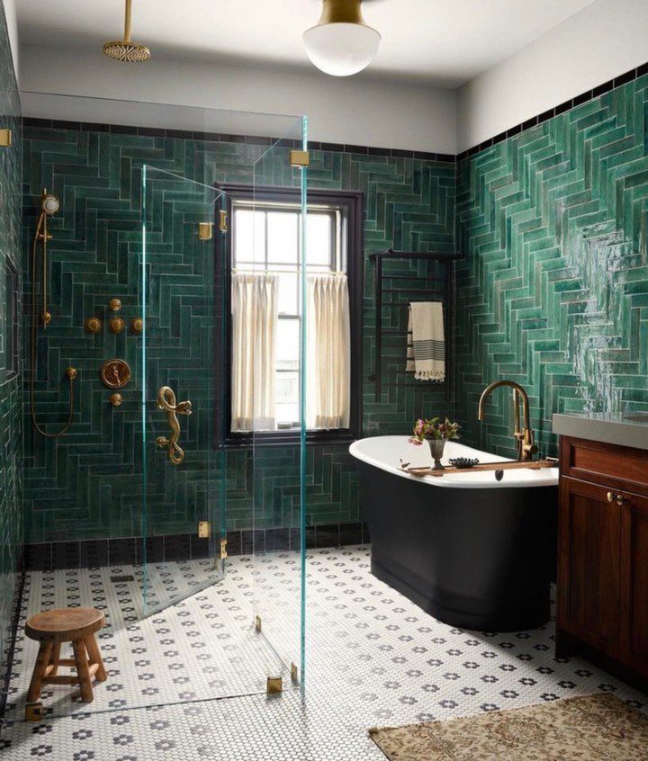 A chic bathroom boasting lustrous deep green tiles with brass fixtures, alongside a freestanding black tub and a herringbone-patterned tile floor.