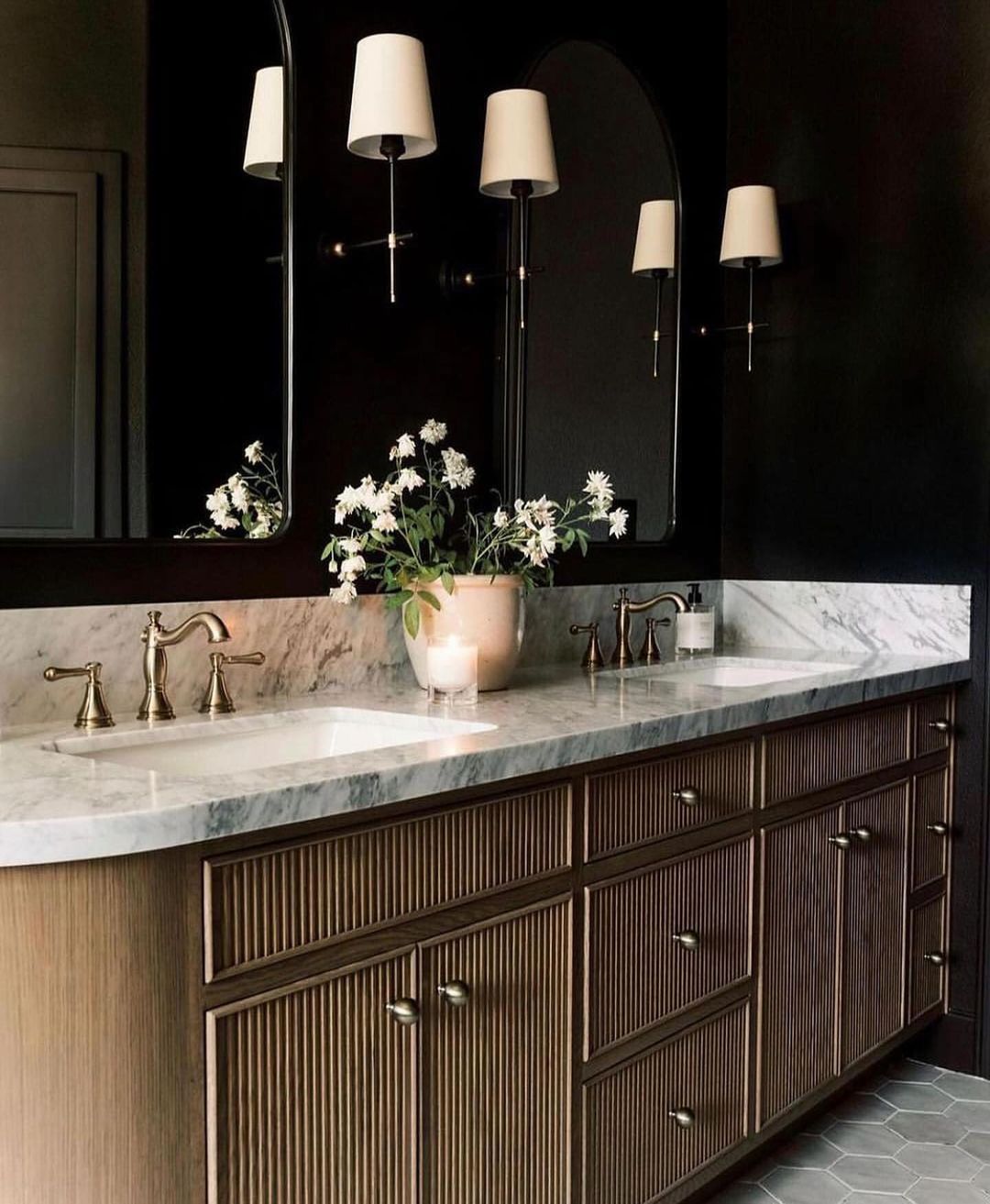 A chic bathroom vanity area featuring dark walls, a marble countertop, and wooden cabinetry with brass fixtures.