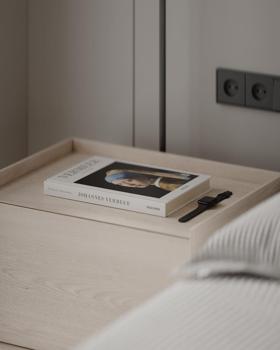 A neatly organized wooden tray featuring a book about Johannes Vermeer and a black wristwatch, suggesting a serene reading nook or bedtime ritual.