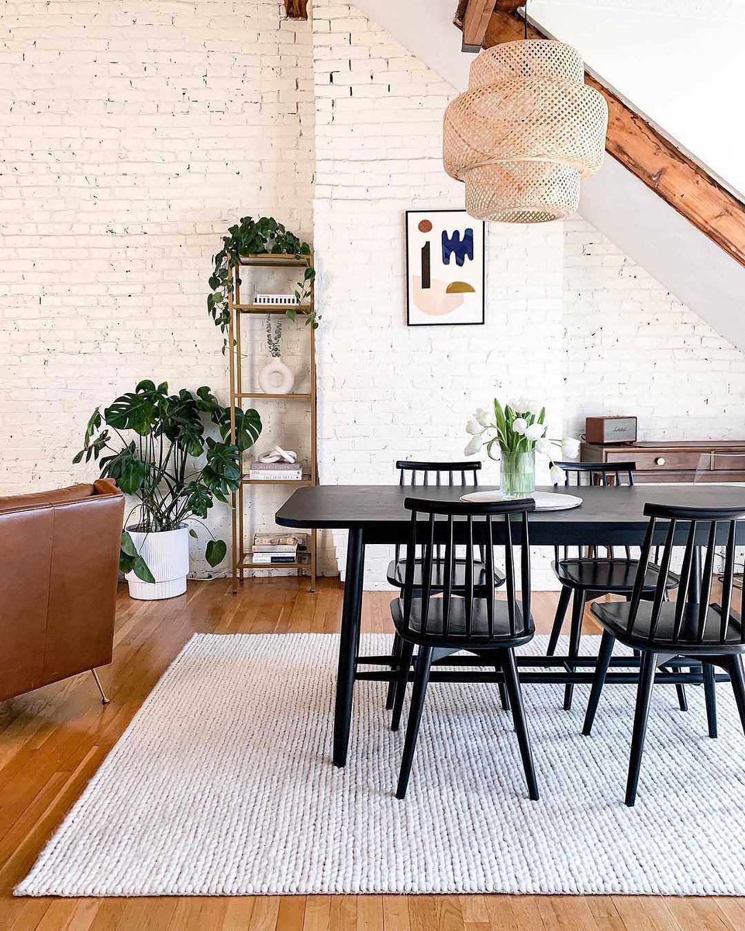 A chic and minimalist dining room featuring a black table with matching chairs set on a textured white rug, against a backdrop of white-painted brick walls and exposed wooden beams.