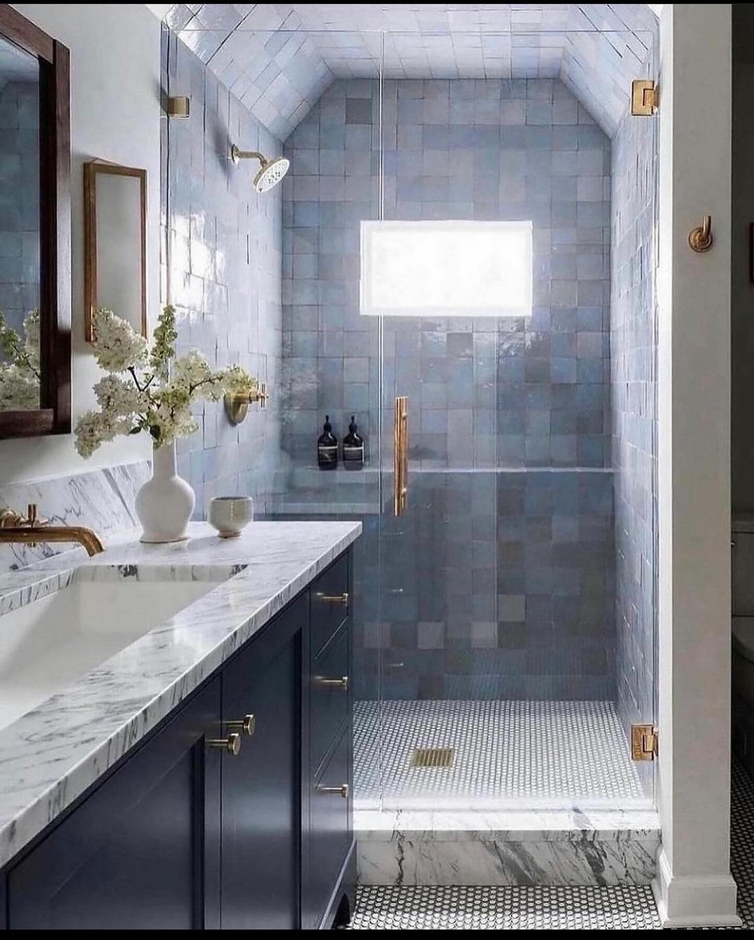 A harmonious blend of textures envelops this bathroom, featuring **gray tiles** in different shades and patterns, a **marble countertop** resting atop a sleek **dark wood vanity** with brass hardware, and a lit mirror casting a glow over the serene space.