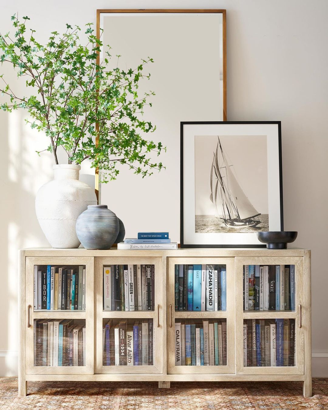 A tastefully arranged living room bookcase featuring a variety of architecture and design books, with a large potted plant on the left and a framed black and white photograph of a sailboat on the right.