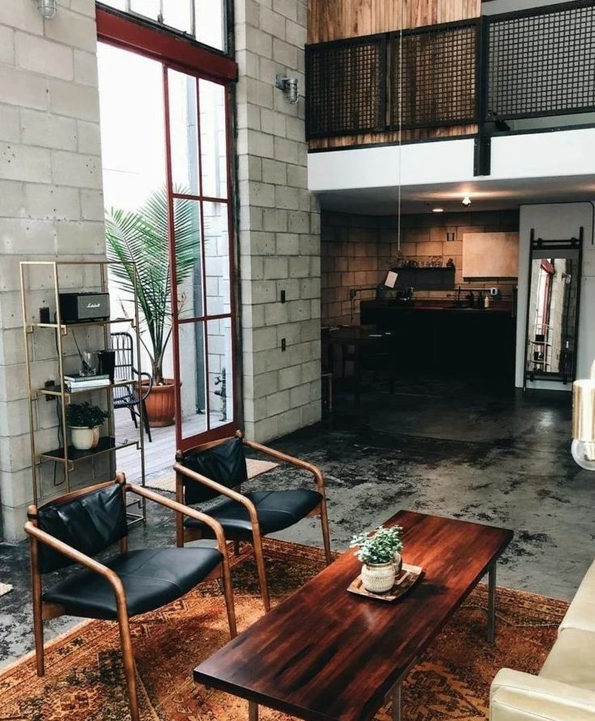 A stylish and minimalist living room with industrial influences featuring exposed concrete walls, a mezzanine with black metal grid railing, warm wooden furniture, and inviting leather chairs