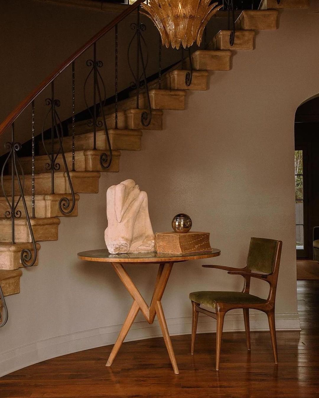 An inviting wooden staircase with ornate iron balustrades against a warm beige wall, accompanied by a bespoke mid-century table and chair set.