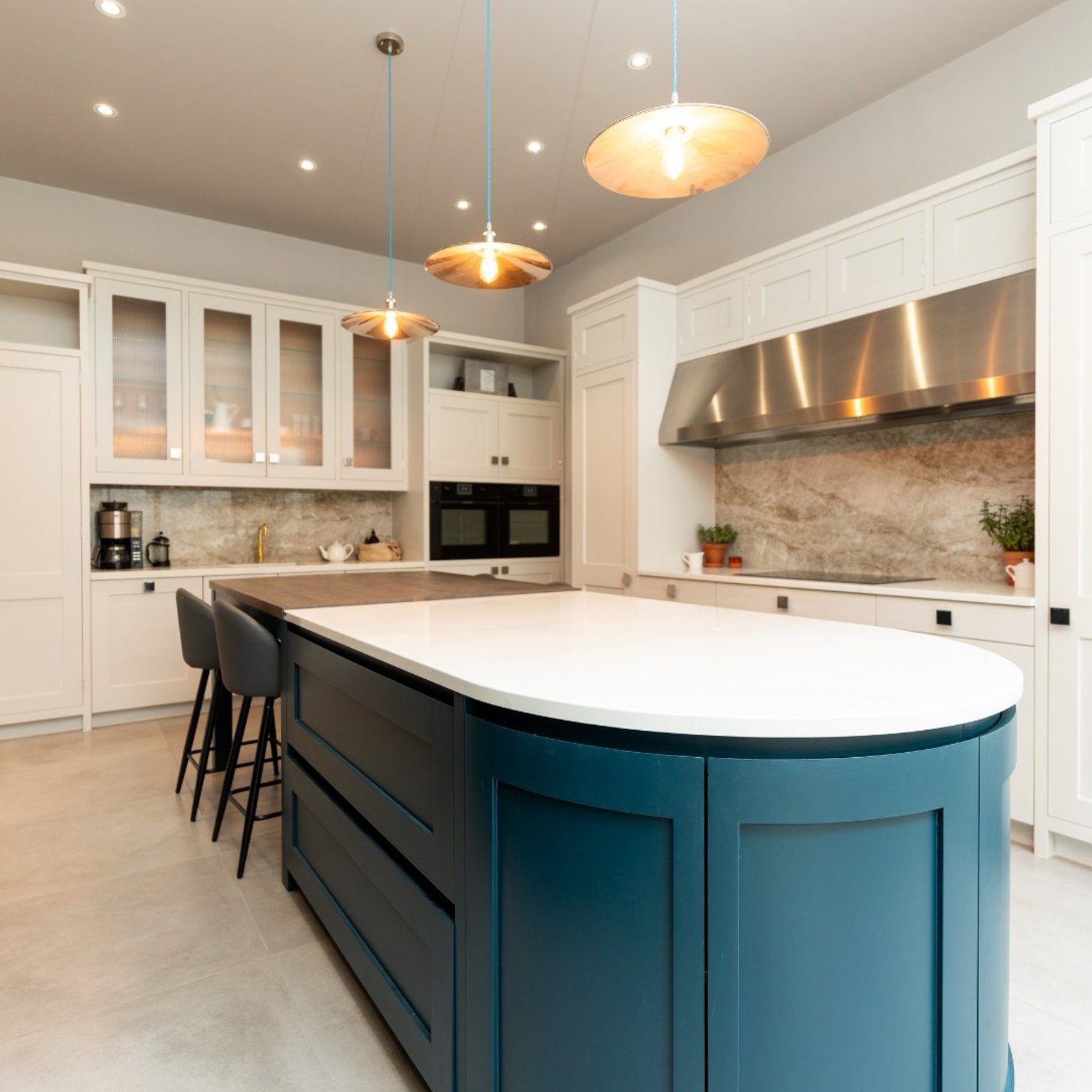 Elegant and spacious kitchen featuring a deep blue island counter with white countertops, white shaker cabinets, built-in stainless steel appliances, and a trio of pendant lights with a warm glow.