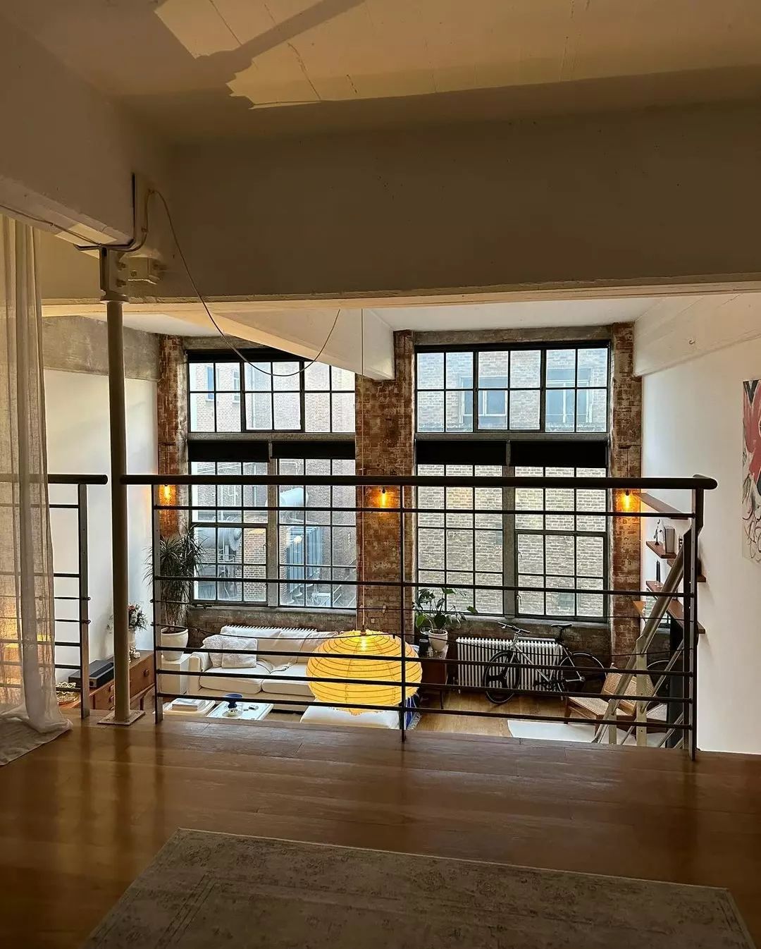 A spacious and urban loft living area featuring exposed brick walls, large windows allowing for ample natural light, and a cozy sitting arrangement with a striking yellow centerpiece.