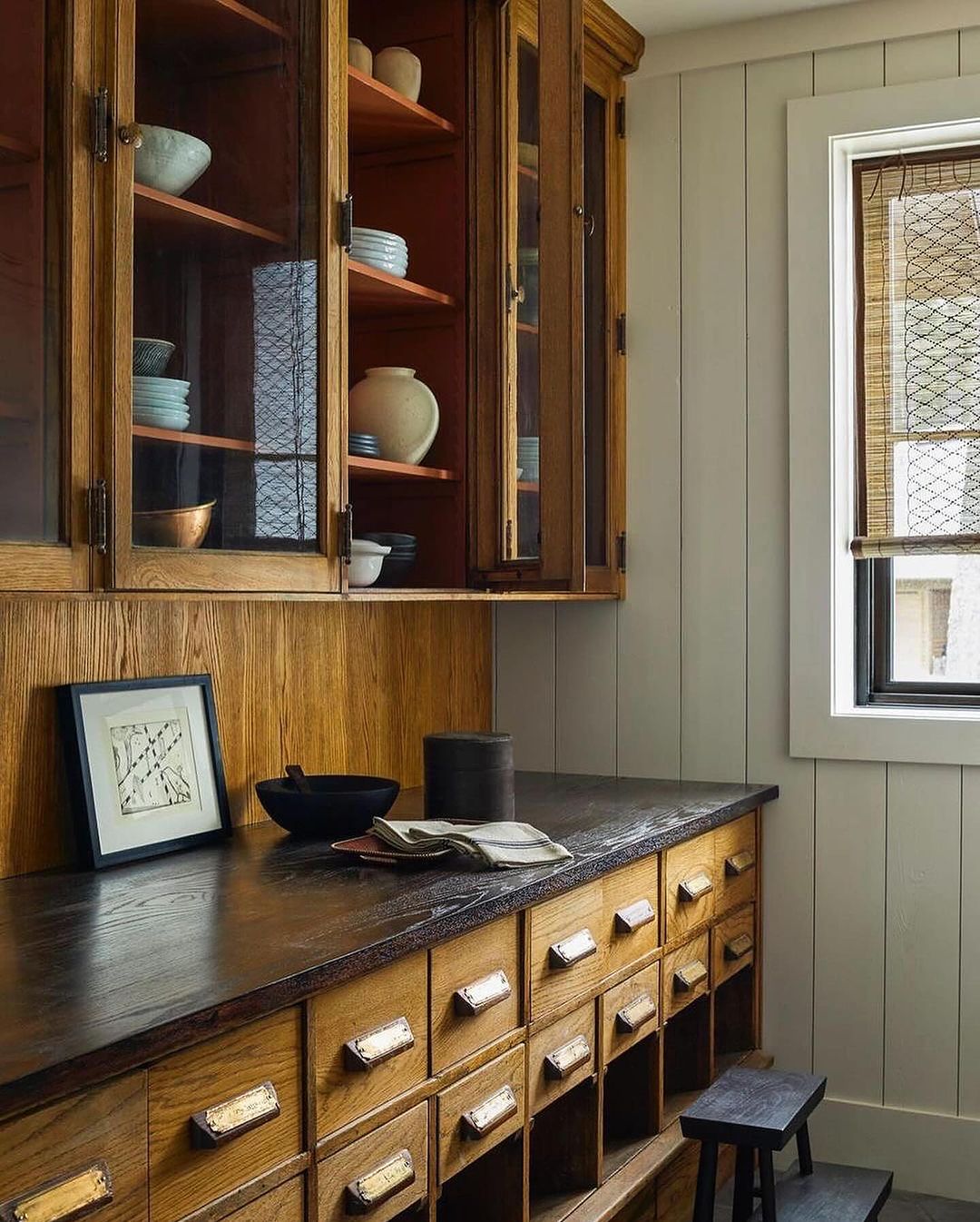 An organized kitchen corner featuring a rustic wooden cabinet with multiple drawers and glass-paneled upper shelves filled with ceramic dishware