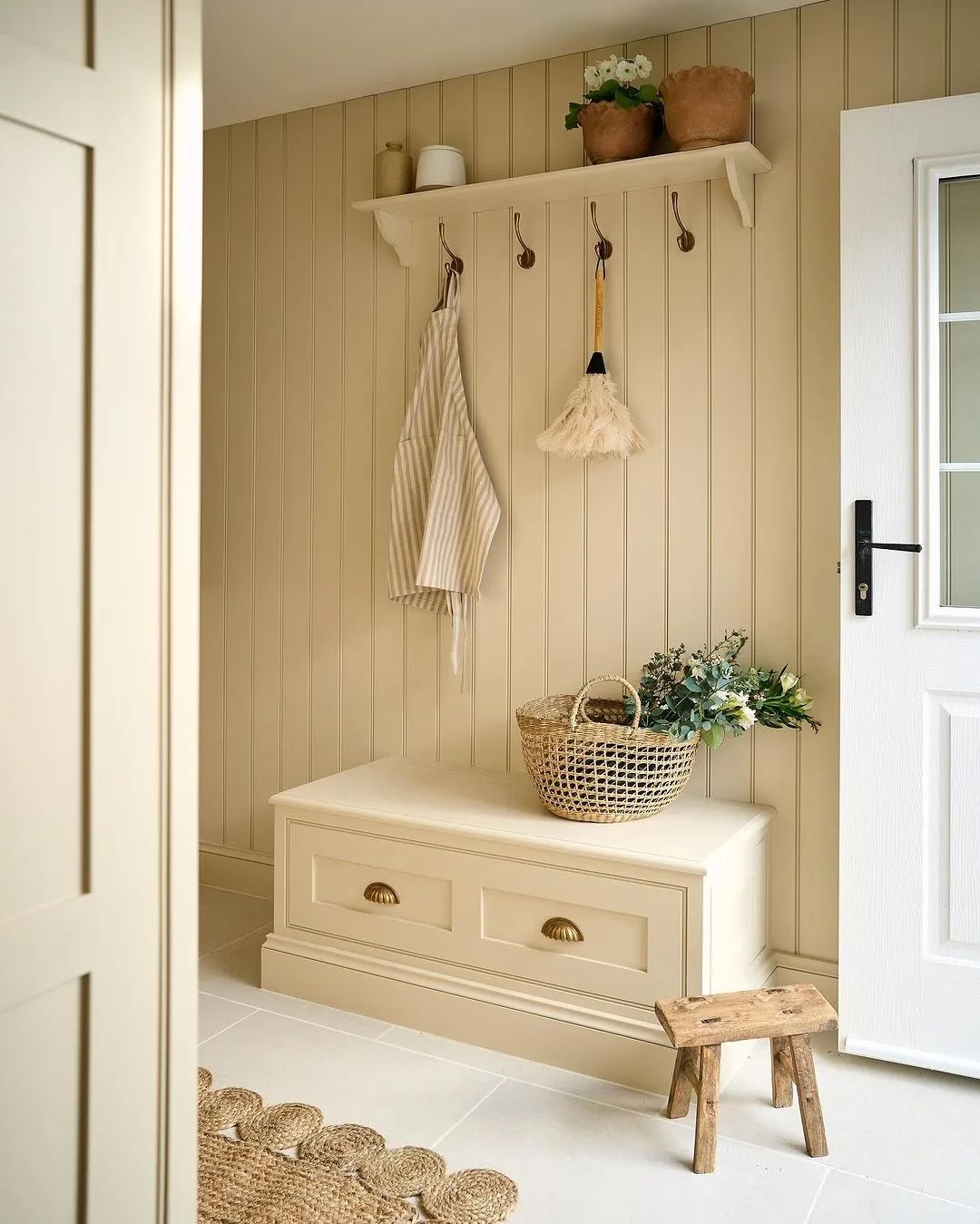 A harmoniously designed mudroom featuring vertical beadboard walls, a built-in cream bench with brass handles, a rustic wooden stool, and a simple shelf displaying pottery and plants.