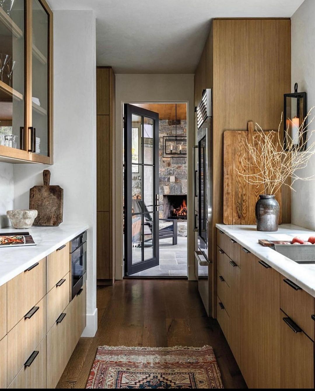 A warm-toned kitchen with sleek wooden cabinetry and white countertops, contrasting with the dark framed glass doors that lead to a cozy fireplace area.